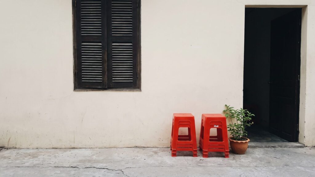 A cream plaster wall with a tall closed wooden shuttered window, a dark open doorway and two stacks of red plastic stools on a cracked concrete floor