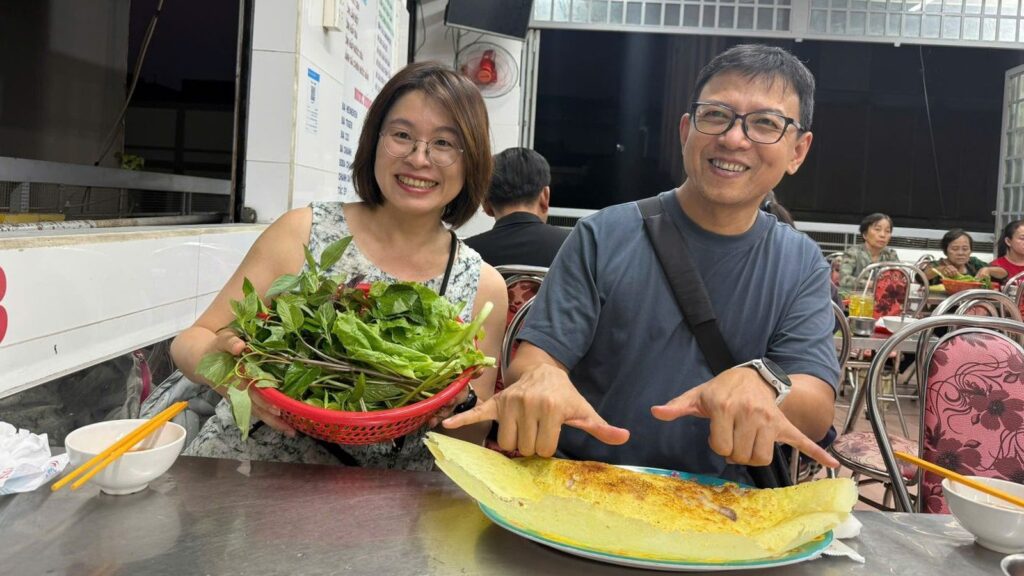 Two travellers sharing a golden banh xeo and a basket of fresh herbs at a steel table in a Saigon street food restaurant