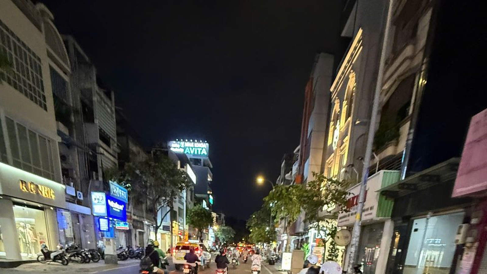 A busy Saigon street after dark seen from the back of a scooter: shop lights, tail lights and riders ahead in the traffic