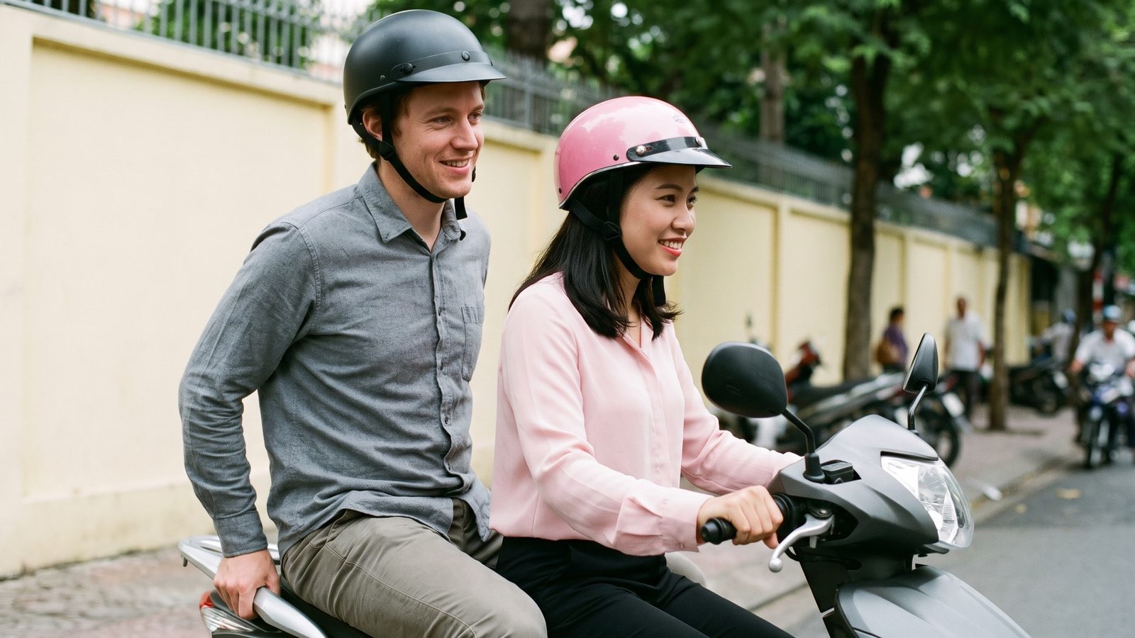A Vietnamese woman guide in a pink helmet drives a scooter while a Western passenger in a black helmet rides behind her holding the rear grab rail on a quiet Saigon street