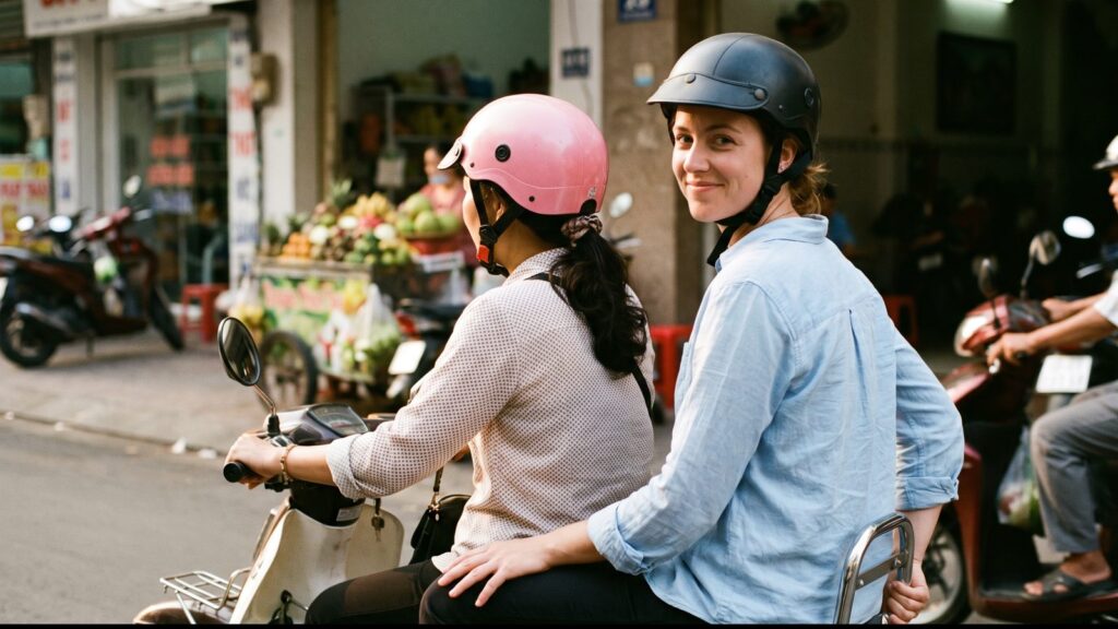 A Vietnamese woman guide in a pink helmet driving a scooter with a Western passenger riding pillion behind her, her hand on the rear grab bar, Saigon street with a fruit stall
