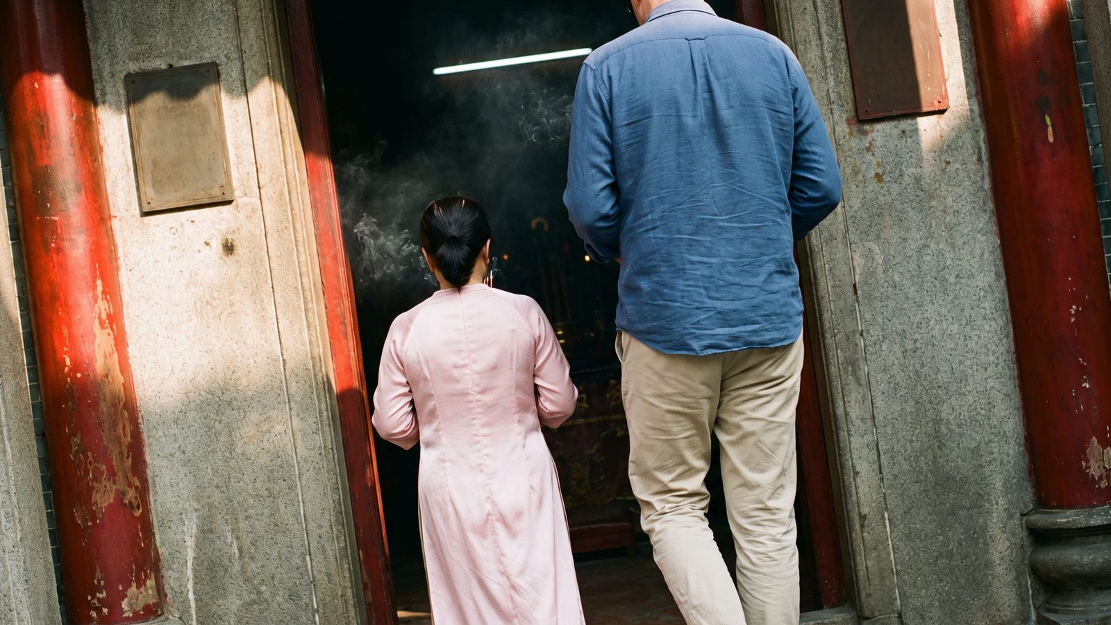 A local woman guide in a pale pink tunic and a visitor seen from behind, walking into the incense-filled entrance of an old Cantonese temple in Cholon, Ho Chi Minh City